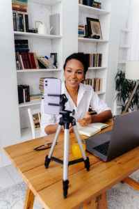 Woman sits at a table with a laptop and cellphone on a tripod.